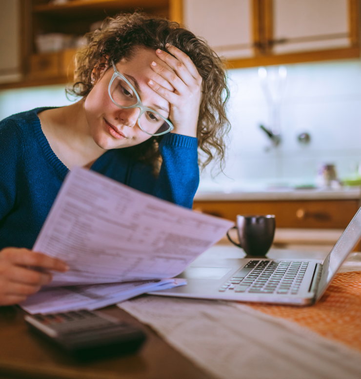 This photo shows a woman in a blue top looking at a bill. She is at a wooden table with her laptop in front of her and a coffee nearby.